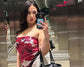 Woman taking a mirror selfie in an elevator wearing a red floral top and white pants, holding a black handbag.